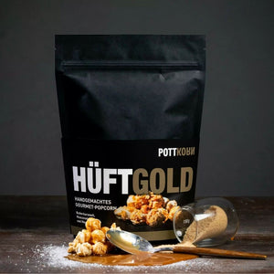 Auf einem Tisch steht eine schwarze Tüte. Auf ihr steht Hüftgold, Pottkorn, handgemachtes Butterkaramell Gourmet Popcorn.
