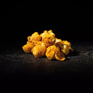 Ein kleiner, kunstvoll arrangierter Haufen goldgelber Popcorn-Stücke liegt auf einer dunklen, leicht strukturierten Holzoberfläche. Der Hintergrund ist tiefschwarz und unbeleuchtet. Ein gezielter Lichtstrahl von rechts oben hebt die unregelmäßige, knusprige Textur und die glänzende Beschichtung des Popcorns hervor, während die linke Seite sanft im Schatten liegt.