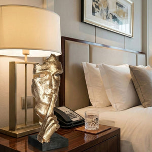 Goldene Skulptur "Silence is Golden" als elegante Dekoration auf dem Nachttisch eines luxuriösen Hotel- oder Schlafzimmers, neben einer Lampe und einem Bett.