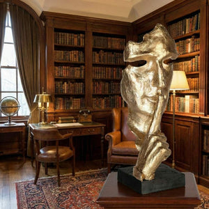 Eine große, goldene Skulptur eines fragmentierten menschlichen Gesichts, das einen Finger an die Lippen legt – die universelle Geste für „Stille“ oder „Psst“. Die Skulptur hat eine raue, strukturierte Oberfläche und steht auf einem schwarzen Sockel im Vordergrund. Der Hintergrund zeigt eine klassische, gemütliche Bibliothek mit deckenhohen Holzregalen voller Bücher, einem braunen Ledersessel, einem antiken Schreibtisch mit Lampe, einem Globus und einem großen Fenster mit Vorhängen.
