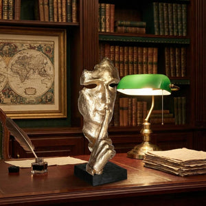 Nahaufnahme der goldenen Skulptur „Silence is Golden“, die ein fragmentiertes Gesicht mit dem Finger auf den Lippen zeigt. Sie steht auf einem dunklen Holzschreibtisch und ist umgeben von einer klassischen Arbeitsatmosphäre: Rechts leuchtet eine grüne Banker-Lampe neben einem Stapel alter Papiere, links ruht ein Federkiel in einem Tintenfass. Im Hintergrund sieht man unscharf dunkle Bücherregale und eine gerahmte, antike Weltkarte an der Wand.