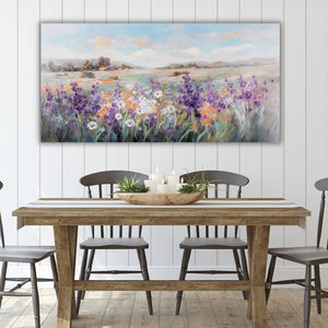 Über einem Tisch mit Holzstühlen hängt an einer weißen Wand ein Bild von einer Sommerwiese in der Toskana. Bunte Blumen stehen auf der Wiese und in der Entfernung sieht man ein typisches Dorf der Toskana oder der Provence.