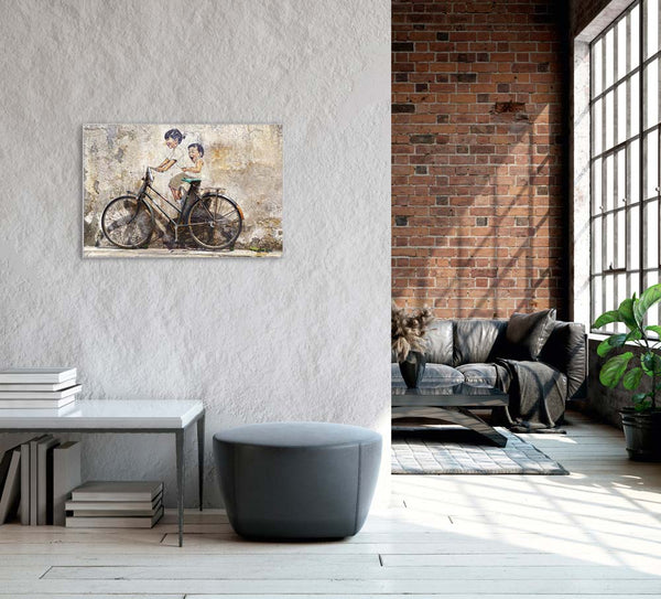An einer grauen Wand hängt eine Bild mit einem Fahrrad. Hinter dem fahrrad sind zwei Kinder an die Hauswand gemalt worden. Dieses Bild hängt an einer grauen Wand in einem Wohnzimmer für Radfahrer.