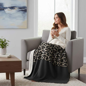 Eine junge Frau sitzt entspannt in einem grauen Sessel, eingehüllt in eine weiche Decke mit Leopardenmuster und einem schwarzen unteren Rand. Sie hält eine Tasse in den Händen und blickt verträumt aus dem Fenster. Im Hintergrund sind ein abstraktes Gemälde an der Wand, eine Zimmerpflanze auf einem Beistelltisch und ein Fenster zu sehen.
