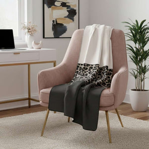 Eine weiche Decke mit einem dreigeteilten Design – oben cremefarben, in der Mitte mit Leopardenmuster und unten dunkelgrau mit feinen Rippen – ist elegant über einen rosafarbenen Sessel mit goldenen Beinen drapiert. Im Hintergrund sind ein weißer Schreibtisch mit einem Laptop und einer Vase mit Blumen, ein abstraktes Gemälde an der Wand und eine große Zimmerpflanze in einem weißen Topf zu sehen. Der Raum wirkt stilvoll und gemütlich.