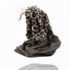 Ein gefaltetes oder schwebendes Leopardenmuster-Decke mit einem beigefarbenen Oberteil und einem dunkelgrauen Unterteil. Die Decke scheint weich zu sein und hat auf der Unterseite eine Steppnaht an den Rändern. Sie ist vor einem hellen, weißen Hintergrund aufgenommen.