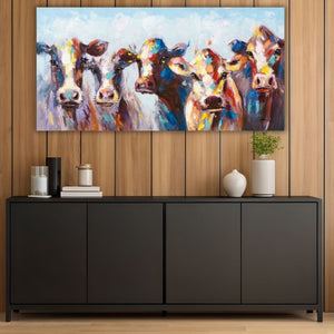 Ein breites, farbenfrohes Gemälde mit fünfKühen hängt an einer Wand mit hellbrauner, vertikaler Holzvertäfelung. Die Kühe sind in kräftigen Farben gemalt und blicken frontal in den Raum. Unter dem Bild steht ein langes, schwarzes Sideboard im modernen Stil, das mit Büchern, Kerzen und Vasen in Weiß und Schwarz dekoriert ist. 